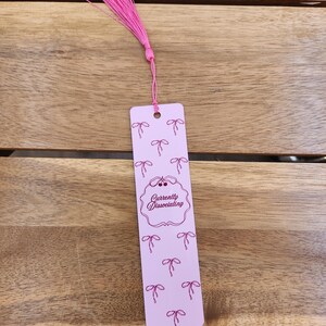 May include: A pink bookmark with a pink tassel. The bookmark has a repeating pattern of pink bows and the text "Currently Dissociating" in a decorative frame. The bookmark is rectangular with a hole at the top.