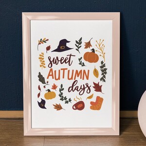 Puede incluir: Un estampado enmarcado con un borde rosa. La obra de arte presenta el texto "sweet AUTUMN days" e ilustraciones de un sombrero de bruja, calabazas, hojas, una taza y un calcetín, todo en colores otoñales. El tema es el otoño.