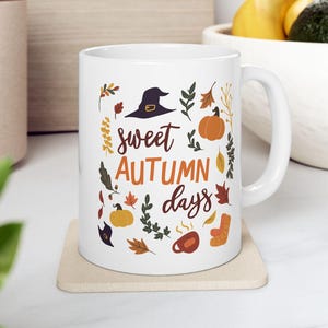 Puede incluir: Taza de cerámica blanca con el texto "sweet AUTUMN days" e ilustraciones otoñales, incluyendo un sombrero de bruja, calabazas y hojas. La taza está sobre un posavasos beige.