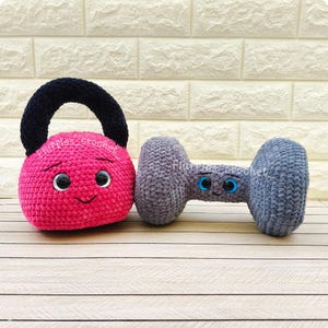 Può includere: Un kettlebell rosa all'uncinetto con manico nero e un manubrio grigio all'uncinetto, entrambi con facce sorridenti e occhi blu. Il testo "Fluffies crochet" è visibile su entrambi gli articoli.