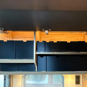 Peut inclure: Intérieur d'une armoire ouverte, révélant des boîtes de rangement noires sur des étagères. L'armoire est en bois clair et possède un dessus sombre. La conception suggère un rangement organisé.