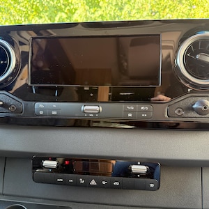 May include: Black dashboard with a central display screen, air vents, and control buttons. The dashboard has a glossy finish and is mounted on a grey surface. The lower section features climate control buttons.