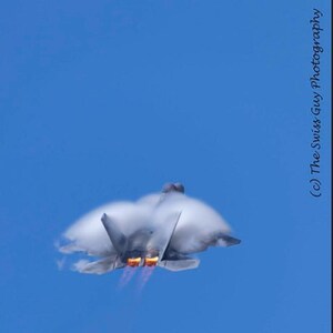 Puede incluir: Un avión de combate gris surca un cielo azul claro, dejando tras de sí una nube de condensación. Los motores del avión emiten llamas naranjas y rojas. La imagen es una fotografía de un avión en vuelo.