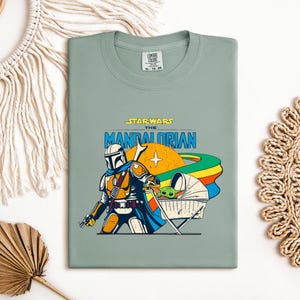 May include: Sage green t-shirt featuring a graphic from the Star Wars series, "The Mandalorian." The design includes the Mandalorian character, a baby Yoda, and the text "STAR WARS THE MANDALORIAN" in a retro style. The shirt is a Comfort Colors brand.