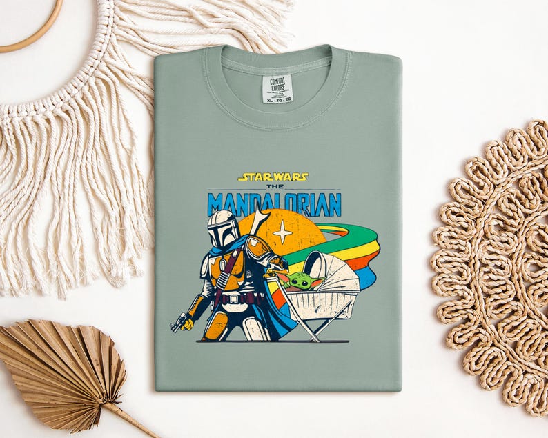 May include: Sage green t-shirt featuring a graphic from the Star Wars series, "The Mandalorian." The design includes the Mandalorian character, a baby Yoda, and the text "STAR WARS THE MANDALORIAN" in a retro style. The shirt is a Comfort Colors brand.