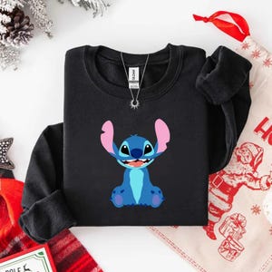 May include: Black crewneck sweatshirt featuring a blue cartoon character with large ears and a smiling expression. The character is set against a black background. A silver necklace is visible at the neckline.