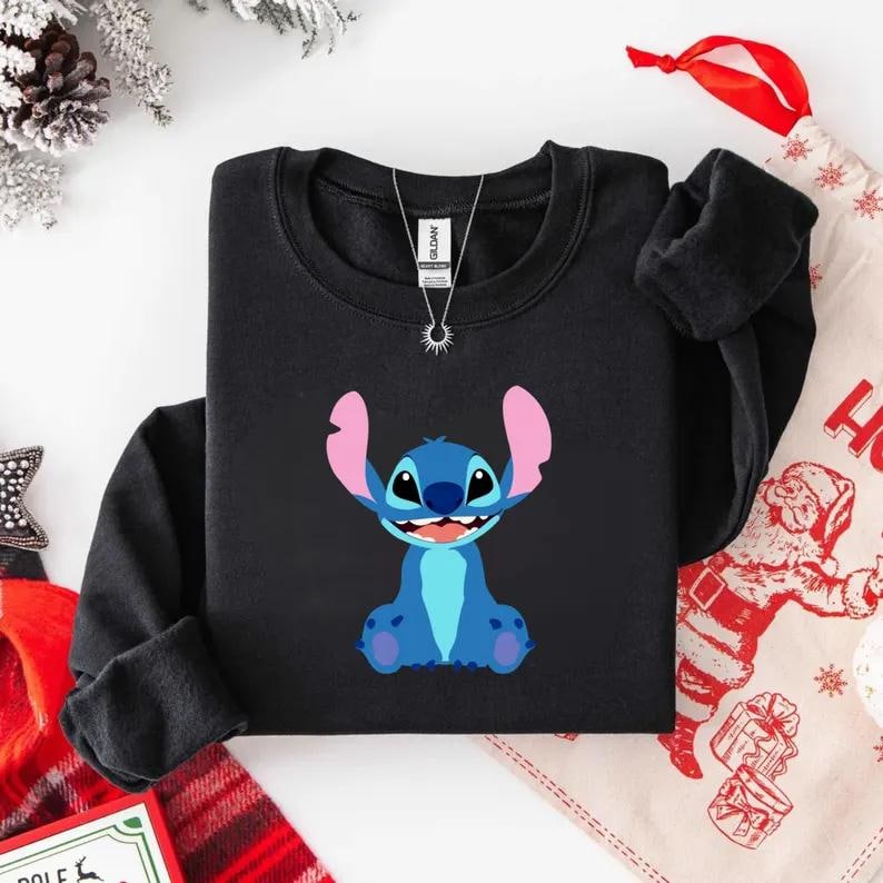 May include: Black crewneck sweatshirt featuring a blue cartoon character with large ears and a smiling expression. The character is set against a black background. A silver necklace is visible at the neckline.