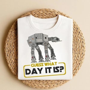 May include: White t-shirt featuring a gray AT-AT walker illustration with the text "GUESS WHAT DAY IT IS?" in a yellow rectangle. The design is centered on the shirt, which is displayed on a woven, circular surface.