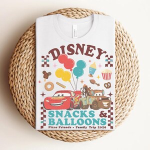 May include: White t-shirt featuring a colorful graphic with the word "Disney" above illustrations of Cars characters, balloons, and snacks. The text "Snacks & Balloons" and "Pixar Friends &bull; Family Trip 2026" are also included.