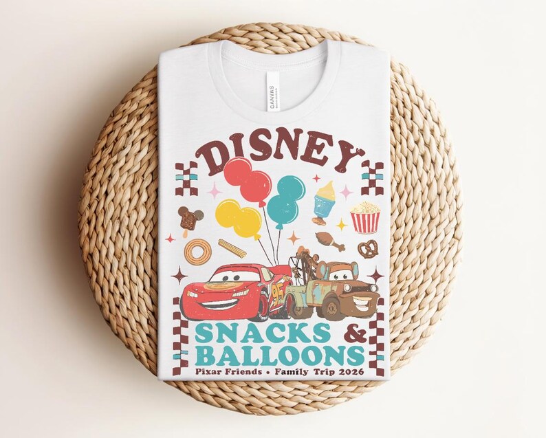 May include: White t-shirt featuring a colorful graphic with the word "Disney" above illustrations of Cars characters, balloons, and snacks. The text "Snacks & Balloons" and "Pixar Friends &bull; Family Trip 2026" are also included.