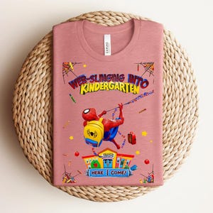 Pu&ograve; includere: T-shirt rosa polveroso con una grafica colorata di Spider-Man che si lancia verso la scuola materna. Il design include il testo "Web-Slinging Into Kindergarten" e "Here I Come!" con illustrazioni scolastiche. La maglietta &egrave; piegata su un tappetino intrecciato.