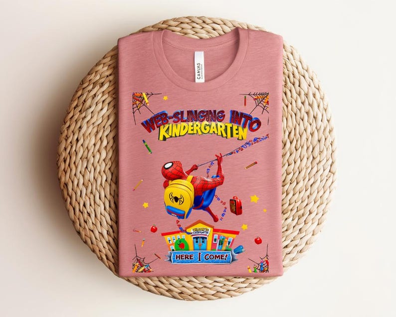 Pu&ograve; includere: T-shirt rosa polveroso con una grafica colorata di Spider-Man che si lancia verso la scuola materna. Il design include il testo "Web-Slinging Into Kindergarten" e "Here I Come!" con illustrazioni scolastiche. La maglietta &egrave; piegata su un tappetino intrecciato.