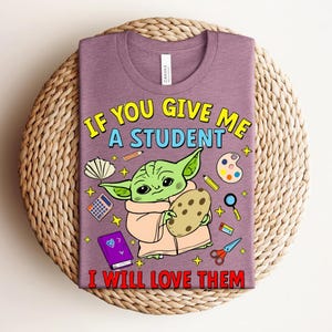 May include: A purple t-shirt with a cartoon image of a green alien-like character holding a cookie, with school supplies. The text says, "IF YOU GIVE ME A STUDENT I WILL LOVE THEM."