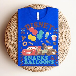 May include: A royal blue t-shirt featuring a colorful graphic design. The design includes the word "DISNEY" in brown letters, balloons, and cartoon cars. The text "SNACKS & BALLOONS" is also visible. The shirt is folded on a woven surface.
