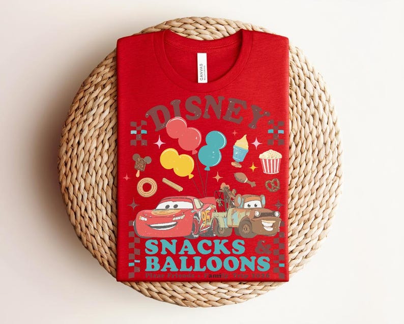 May include: Red t-shirt featuring a graphic of Lightning McQueen and Mater from the movie Cars, along with balloons and snacks. The shirt has the word "Disney" above the graphic and "Snacks Balloons" below. The shirt is folded on a woven mat.
