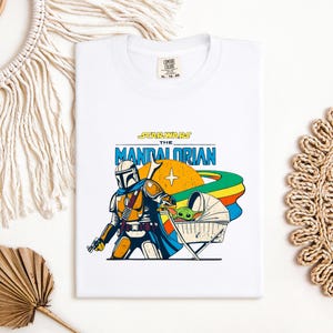 May include: White t-shirt featuring a colorful graphic of The Mandalorian and Grogu from Star Wars. The design includes the text "STAR WARS THE MANDALORIAN" in blue and yellow, with a retro-style illustration of the characters and a rainbow.