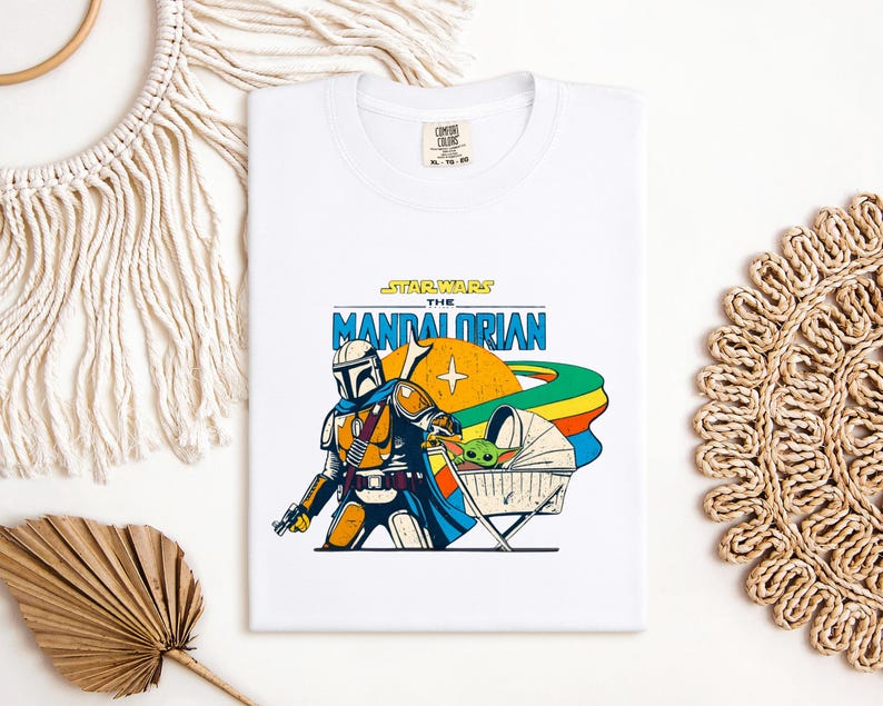 May include: White t-shirt featuring a colorful graphic of The Mandalorian and Grogu from Star Wars. The design includes the text "STAR WARS THE MANDALORIAN" in blue and yellow, with a retro-style illustration of the characters and a rainbow.
