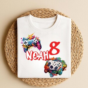 May include: White t-shirt featuring colorful illustrations of two video game controllers. The name "NOAH" is printed in red, along with the number "8". The controllers have vibrant accents of blue, green, yellow, and pink.