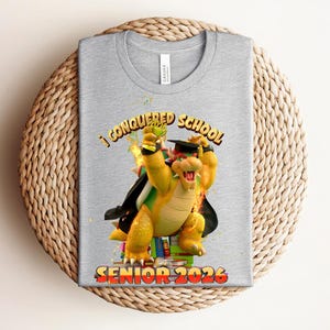 May include: A light gray t-shirt featuring a cartoon Bowser character in a graduation cap, holding a trophy. The text "I CONQUERED SCHOOL SENIOR 2026" is printed in yellow and red. The shirt is made of a soft material.