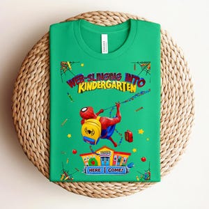Pu&ograve; includere: Una maglietta verde con una grafica colorata di Spider-Man che si dondola con uno zaino e un portapranzo. Il testo "WEB-SLINGING INTO KINDERGARTEN" &egrave; visualizzato sopra il personaggio, con "HERE I COME!" sotto.