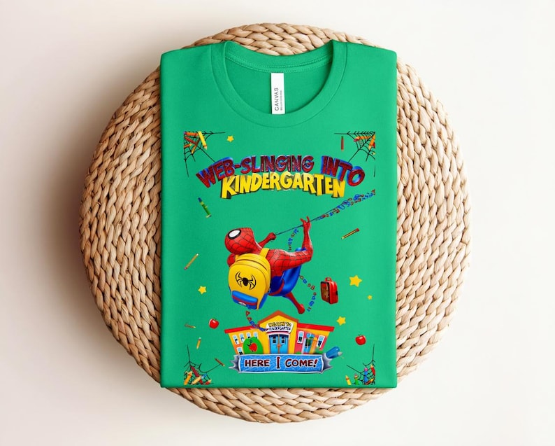 Pu&ograve; includere: Una maglietta verde con una grafica colorata di Spider-Man che si dondola con uno zaino e un portapranzo. Il testo "WEB-SLINGING INTO KINDERGARTEN" &egrave; visualizzato sopra il personaggio, con "HERE I COME!" sotto.