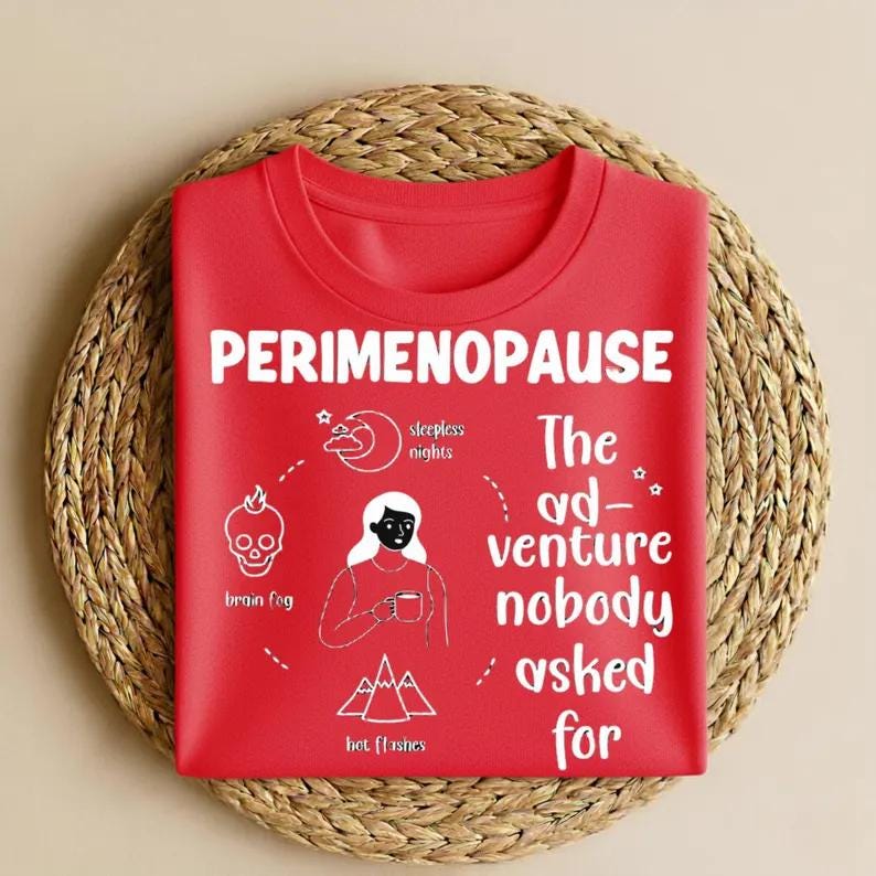 Op de afbeelding: Een rood T-shirt met ronde hals met witte tekst en illustraties. Het shirt leest "PERIMENOPAUSE" en "The ad-venture nobody asked for." Illustraties zijn onder meer een schedel, maan, vrouw en bergen, met labels als "brain fog" en "hot flashes."