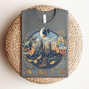 May include: Dark gray t-shirt featuring a colorful graphic with the text "UNIVERSAL STUDIOS." The design includes a dinosaur, a minion, a castle, and other iconic imagery from popular movies and attractions.
