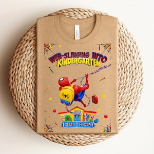 Pu&ograve; includere: T-shirt color cachi con grafica colorata di Spider-Man che si dondola, con la scritta "Web-Slinging Into Kindergarten". Il design include una scuola e la frase "Here I Come!" in un carattere giocoso.