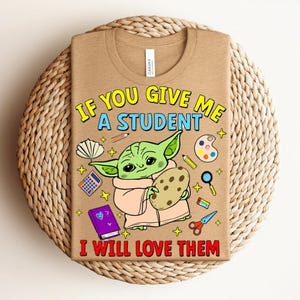 May include: A beige t-shirt with the text "IF YOU GIVE ME A STUDENT I WILL LOVE THEM" and a cartoon image of a green alien holding a cookie. The shirt also features school-related graphics.