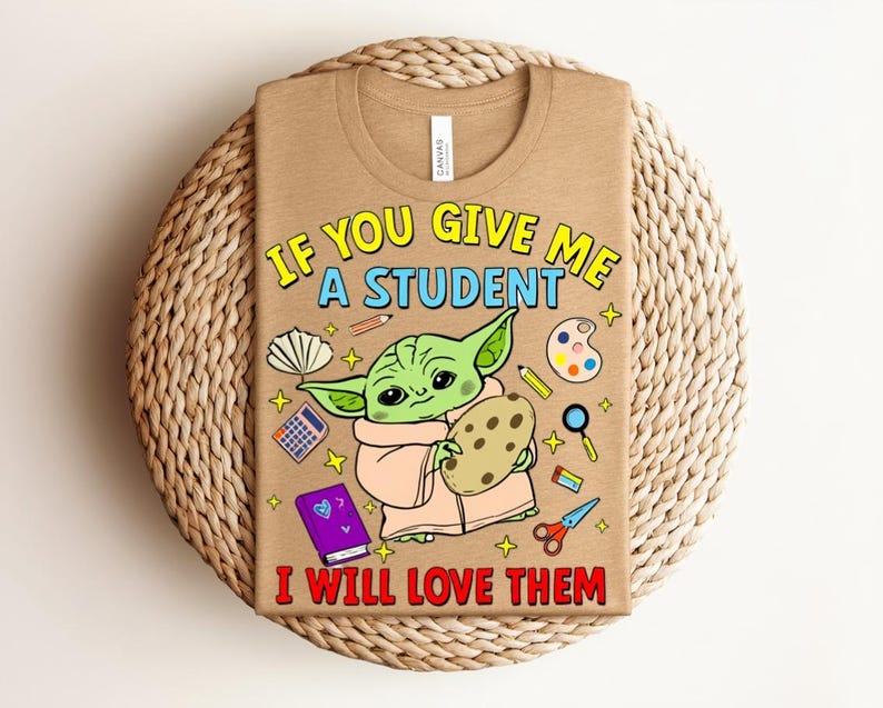 May include: A beige t-shirt with the text "IF YOU GIVE ME A STUDENT I WILL LOVE THEM" and a cartoon image of a green alien holding a cookie. The shirt also features school-related graphics.