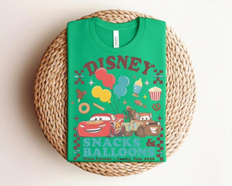 May include: Green t-shirt with a graphic featuring the word "DISNEY" in red, balloons, and cartoon cars. The text "SNACKS & BALLOONS" is below the cars. The shirt is on a woven, circular surface.