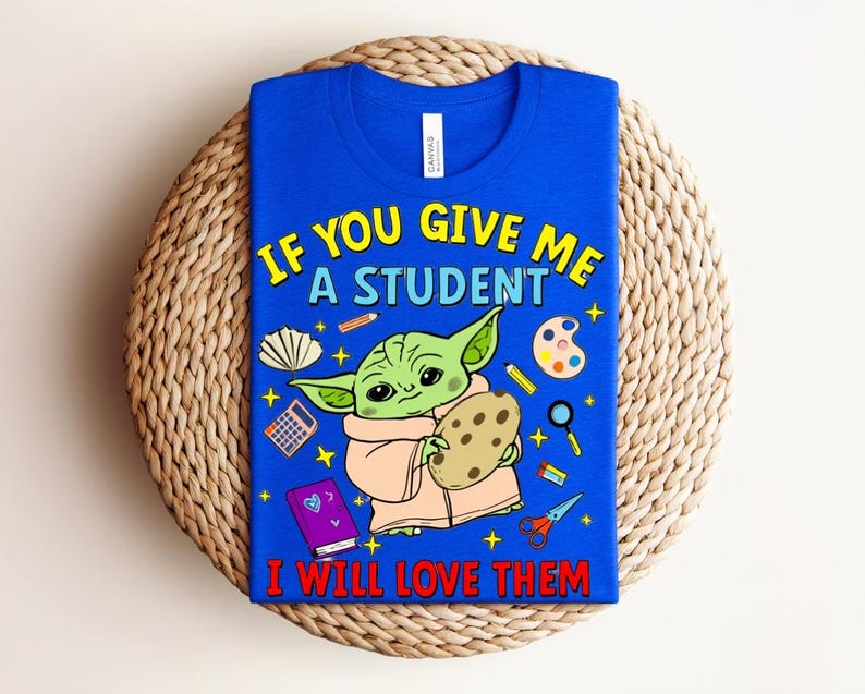 May include: Royal blue t-shirt with the text "IF YOU GIVE ME A STUDENT I WILL LOVE THEM". The graphic features a green alien-like character holding a cookie, surrounded by school-themed illustrations. The shirt is displayed on a woven mat.