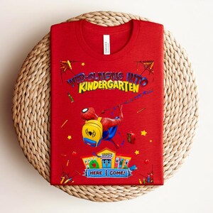 Pu&ograve; includere: Una maglietta rossa con una grafica di Spider-Man e la scritta "Web-Slinging Into Kindergarten". Il design include un Spider-Man dei cartoni animati, uno zaino giallo e la frase "Here I Come!" in un banner colorato.