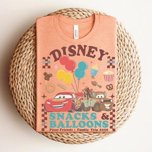 May include: Orange t-shirt featuring a Disney design with the text "Disney Snacks & Balloons". The graphic includes cartoon cars, balloons, and food icons. The shirt is on a woven, circular surface.