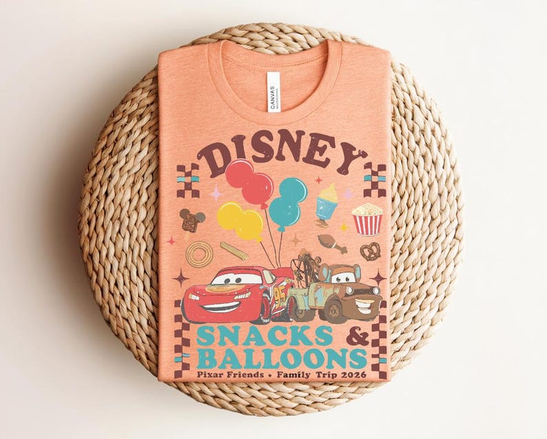 May include: Orange t-shirt featuring a Disney design with the text "Disney Snacks & Balloons". The graphic includes cartoon cars, balloons, and food icons. The shirt is on a woven, circular surface.