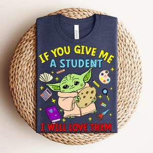 May include: A navy blue t-shirt with a cartoon character holding a cookie, surrounded by school-themed illustrations. The text reads "IF YOU GIVE ME A STUDENT I WILL LOVE THEM" in yellow and red.