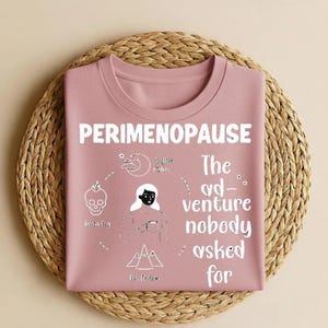 Op de afbeelding: Een stoffig roze t-shirt met het woord "PERIMENOPAUSE" in witte blokletters. Het shirt heeft een afbeelding van een vrouw en symbolen die symptomen vertegenwoordigen zoals hersenmist, slapeloze nachten en opvliegers. De tekst "The ad-venture nobody asked for" wordt ook weergegeven.