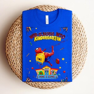 Pu&ograve; includere: Una maglietta blu con la scritta "WEB-SLINGING INTO KINDERGARTEN" e una grafica di Spider-Man. Il disegno include un edificio scolastico, uno zaino e altri elementi scolastici. &Egrave; visibile anche la scritta "HERE I COME!".