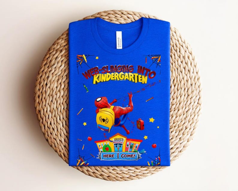 Pu&ograve; includere: Una maglietta blu con la scritta "WEB-SLINGING INTO KINDERGARTEN" e una grafica di Spider-Man. Il disegno include un edificio scolastico, uno zaino e altri elementi scolastici. &Egrave; visibile anche la scritta "HERE I COME!".
