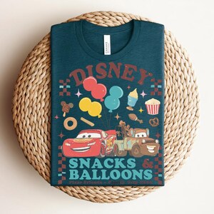 May include: Teal t-shirt with a graphic featuring the word "Disney" above colorful balloons, snacks, and cartoon cars. The text "Snacks & Balloons" is below the cars. The shirt is displayed on a woven, circular surface.
