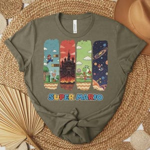 May include: Olive green t-shirt featuring the text "SUPER MARIO" in blue and yellow. The shirt has four vertical rectangular graphics with scenes from the Super Mario video game, including Mario, a castle, Yoshi, and a galaxy.