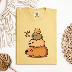 May include: Yellow t-shirt featuring a stack of cartoon capybaras, with the text "STACK OF CAPY" in black. The capybaras are in shades of brown and green. The shirt is folded on a white surface.