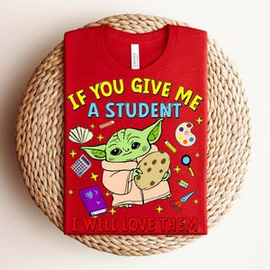 May include: Red t-shirt featuring a green alien-like character holding a cookie, with school supplies illustrations. The text says "IF YOU GIVE ME A STUDENT I WILL LOVE THEM" in yellow, blue, and white.