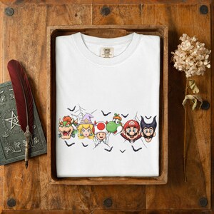 May include: A white t-shirt featuring a graphic print of Bowser, Princess Peach in a witch hat, Toad, Yoshi with a pumpkin, Mario, and Luigi with horns, all surrounded by black spiderwebs and small bat silhouettes across the chest area.