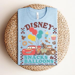May include: Light blue t-shirt with a colorful graphic featuring the word "Disney" above a design of balloons, food, and cartoon cars. The text "Snacks & Balloons" and "Pixar Friends &bull; Family Trip 2026" are also printed on the shirt.