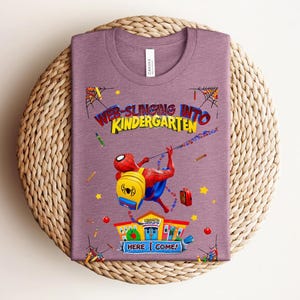 Pu&ograve; includere: Una maglietta malva con una grafica colorata di Spider-Man che entra a scuola. Il testo recita "Web-Slinging Into Kindergarten" con le parole "Here Come!". Il design include un edificio scolastico, matite e stelle.