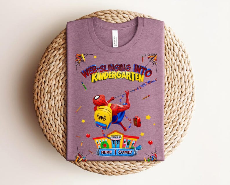 Pu&ograve; includere: Una maglietta malva con una grafica colorata di Spider-Man che entra a scuola. Il testo recita "Web-Slinging Into Kindergarten" con le parole "Here Come!". Il design include un edificio scolastico, matite e stelle.