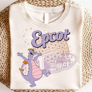 May include: Cream-colored t-shirt featuring the word "Epcot" in purple, with a cartoon dragon character and a stylized architectural design in the background. The design includes stars and a futuristic cityscape.
