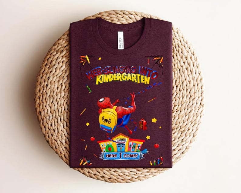 Pu&ograve; includere: Una maglietta bordeaux con una grafica colorata di Spider-Man che va a scuola. Il testo recita "Web-Slinging Into Kindergarten" con "Here I Come!" sotto l'illustrazione della scuola. La maglietta ha un girocollo.