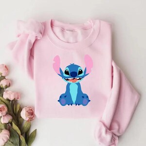 May include: A light pink sweatshirt featuring a cartoon illustration of Stitch from Lilo & Stitch. The character is blue with large ears and a wide smile. The sweatshirt has long sleeves and a crew neck.